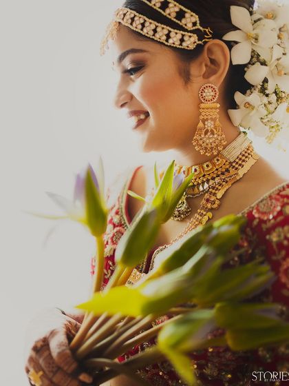 A high-key portrait of a smiling bride holding lotus buds. Her joyful expression, traditional gold jewelry, and the gajra in her hair are all highlighted against a bright, clean background.