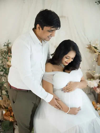 A classic and loving couple's portrait in an all-white setting. The husband stands behind his seated wife, his hands gently on her shoulders, creating a sense of unity and support.