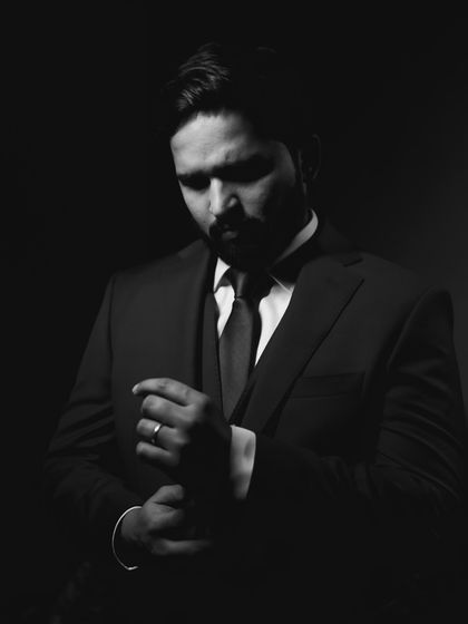 A moody and sophisticated black and white portrait. The shadows and focused light on the groom adjusting his cufflink create a cinematic and timeless feel.