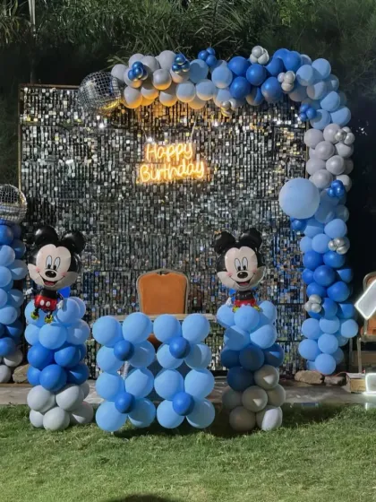 An outdoor Mickey Mouse first birthday party with a sequin backdrop, blue and silver balloon arch, and character cutouts.