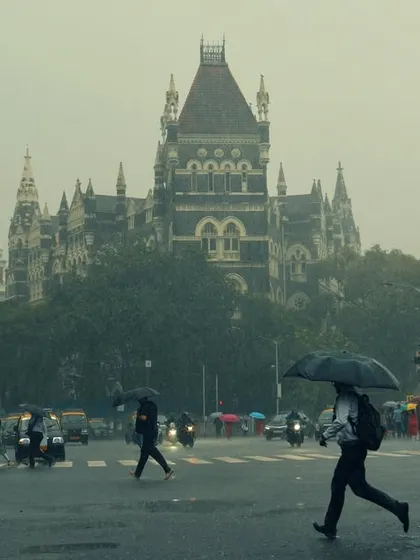 Pedestrians cross a wet street with the historic High Court building looming in the background. The image captures the daily rhythm of Mumbai life that continues, beautifully and stoically, even in the middle of a downpour.