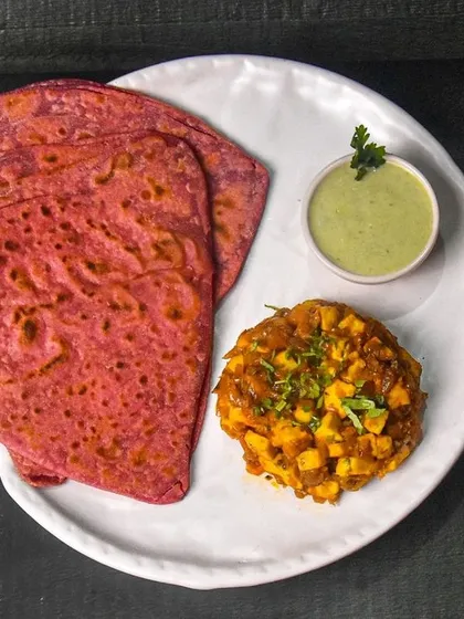 A unique Indian meal of beetroot paratha served with a savory paneer masala and mint chutney. The dark background makes the vibrant pink of the paratha stand out.