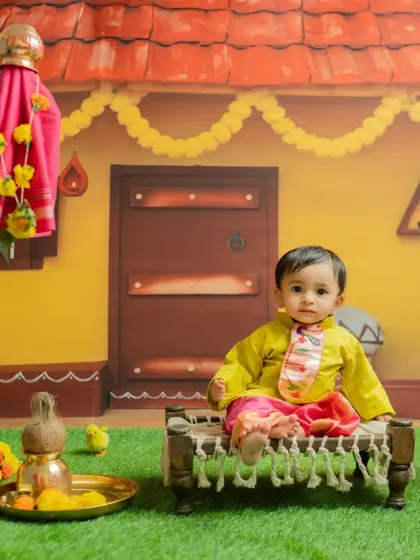 Sitting on a miniature 'charpai' or traditional cot, this baby looks adorable in the Gudi Padwa setup, which features a village house backdrop and festive marigold flowers.