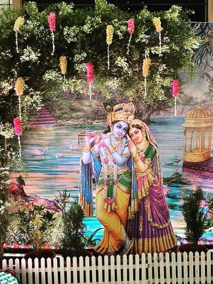 A beautiful photo booth with a Radha Krishna theme. The backdrop is a serene painting of the divine couple, framed by a real tree decorated with floral hangings.
