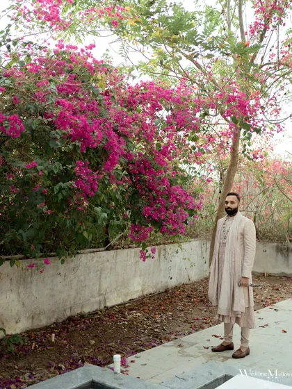 The groom stands against a vibrant wall of pink bougainvillea, creating a striking contrast with his elegant attire. A beautiful and artistic groom portrait.