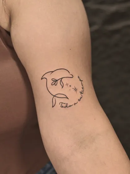 A playful and simple single-line dolphin tattoo. The design incorporates a flower and sparkling stars, with a delicate script that reads "Todo a su tiempo" (Everything in its own time).