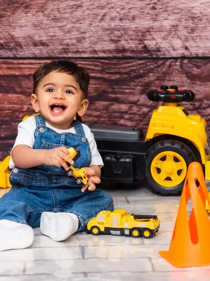 A fun and creative construction-themed photoshoot with a smiling baby boy in denim overalls, surrounded by toy trucks and traffic cones.