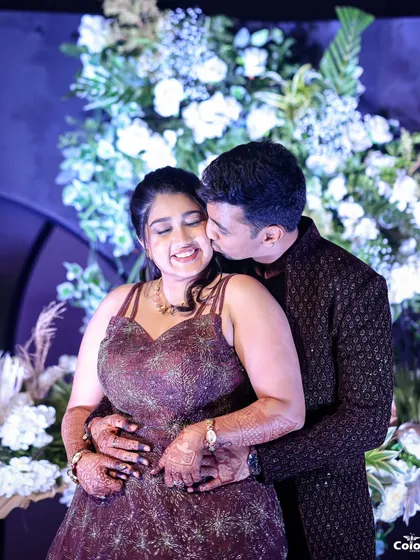 A sweet, candid moment as the groom kisses the bride's cheek against a beautiful floral backdrop.