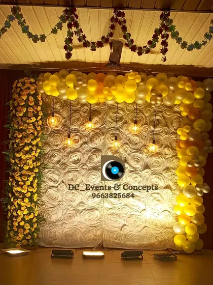 A simple yet elegant birthday backdrop. The design uses a textured white rose wall framed by yellow balloons and flowers, with hanging Edison bulbs adding a warm and celebratory glow.