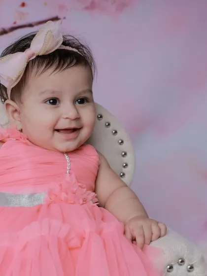 A classic portrait of a smiling baby girl in a beautiful pink dress against a soft, floral backdrop. These sitter session photos are perfect for capturing that milestone when your child can sit up and their personality shines.