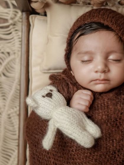 A close-up portrait of the baby in the brown swaddle and bonnet, highlighting their delicate features. The earthy color palette is both modern and timeless.