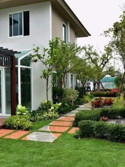 A charming garden path made of brick and stone pavers meanders through a lushly planted residential yard. The design uses a variety of shrubs and trees to create depth and year-round interest.
