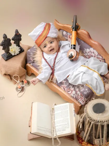 A creative overhead shot of a baby dressed as a Varkari, lying on a miniature cot. The props, including a tabla, a veena, and a holy book, are arranged around him to create a complete narrative.