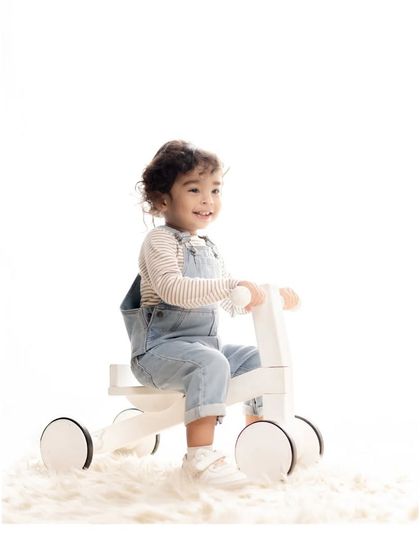 A toddler beams with a huge smile while sitting on a modern, minimalist wooden tricycle. The clean white background and soft rug ensure that his infectious joy is the absolute center of attention.