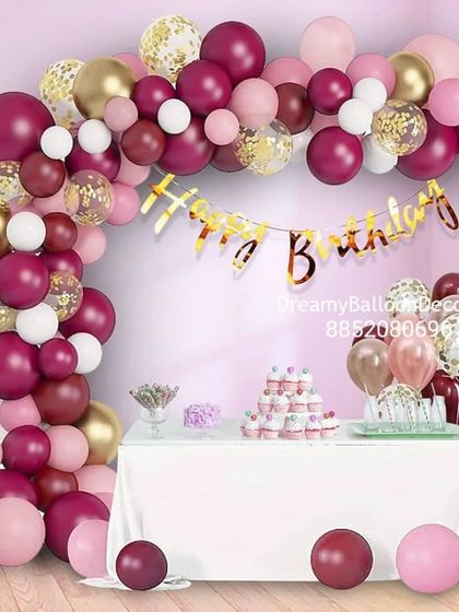 A beautiful dessert table setup with a balloon arch in shades of burgundy, pink, and gold. The confetti balloons and 'Happy Birthday' banner add a touch of elegance.