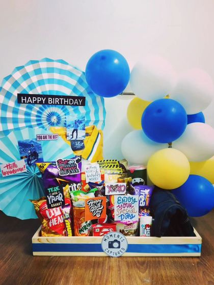 A cheerful blue and yellow themed birthday hamper. This gift is packed with an assortment of snacks and decorated with balloons and paper fans, making it a bright and happy surprise.