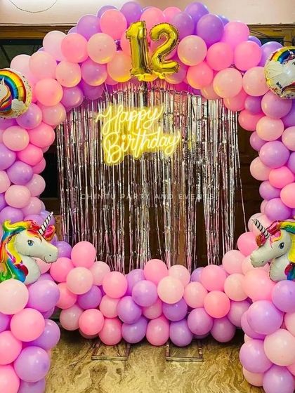 This unicorn-themed birthday setup features a full balloon arch with unicorn head foil balloons and a silver fringe curtain backdrop. A 'Happy Birthday' neon sign and a number '12' foil balloon mark the occasion.