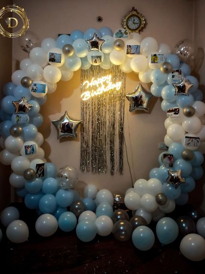 A personalized birthday arch perfect for a home celebration. This blue and white balloon garland is decorated with small photos of the birthday person, star-shaped foil balloons, and a neon sign for a truly custom and heartfelt touch.