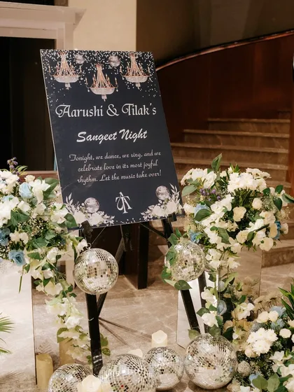 A chic welcome sign for a Sangeet night, complete with sparkling disco balls and elegant white and blue floral arrangements.