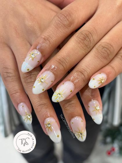 A celestial-themed holiday set. These nails feature a soft white ombré base with hand-painted gold starbursts, each centered with a sparkling crystal.