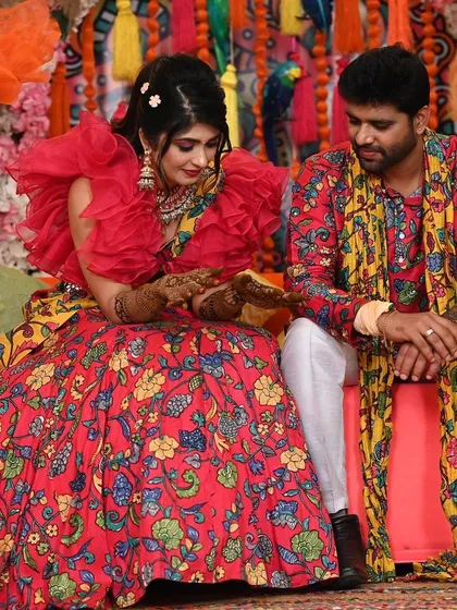 The couple admiring the bride's beautiful mehendi design. The background is filled with colorful tassels, pampas grass, and floral arrangements.