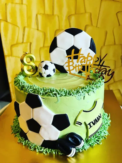 Score a goal with a football-themed birthday cake. This design features a gooey chocolate cake covered in fresh whipped cream "grass" and decorated with fondant footballs and other accents, perfect for any soccer enthusiast.