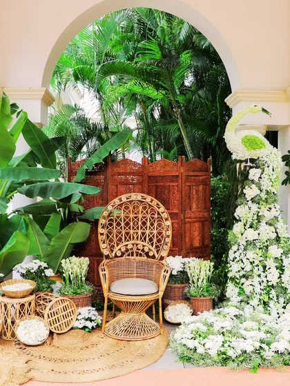 A cozy and thematic seating area for the bride. The wicker peacock chair, surrounded by lush greenery and a peacock made of white flowers, creates a perfect photo spot that is both rustic and elegant.