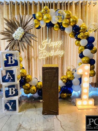 A classic and elegant first birthday decoration in blue, gold, and white. This setup includes a balloon arch, a 'Happy Birthday' neon sign, 'BABY' blocks, and a light-up number '1', perfect for a baby boy's celebration.