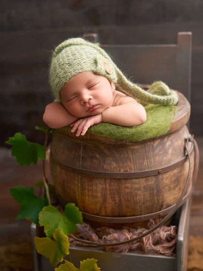 A baby in a green knitted pixie hat rests in a wooden bucket, with green vines adding to the natural, woodland theme.