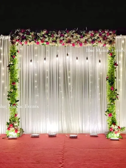 An elegant stage backdrop with a white fairy light curtain and pillars of greenery and flowers. The hanging Edison bulbs add a touch of industrial chic to the otherwise classic design.