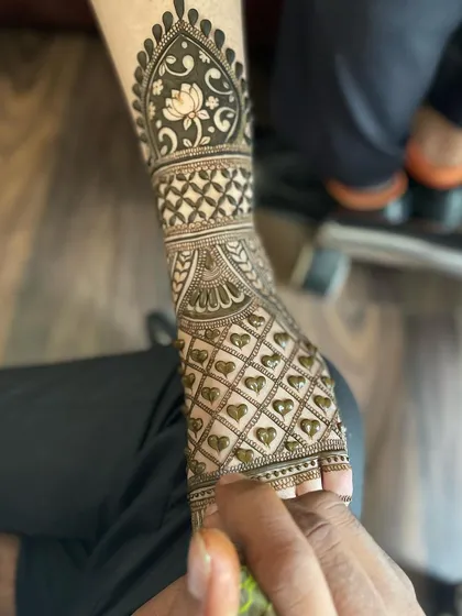 A close-up of a foot mehandi being applied, showing the heart-filled jaali pattern and the lotus motif band on the leg.