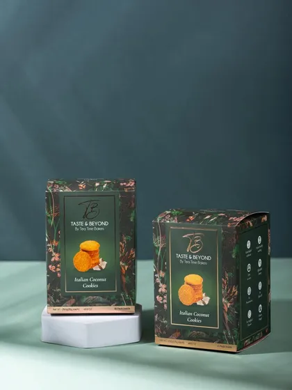 A product shot of Taste & Beyond's Indian Coconut Cookies, using a dark green background and clean geometric props. The lighting highlights the elegant packaging design.