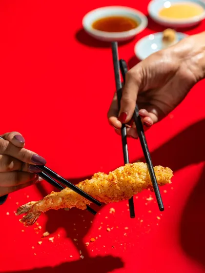 A dynamic shot of two people interacting with crispy shrimp tempura against a bold red background for Harajuku Tokyo Cafe.