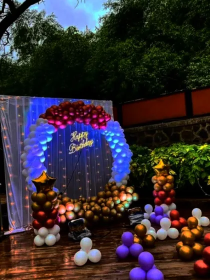 An outdoor birthday backdrop set against lush greenery. The circular balloon arrangement is lit from within, making the neon sign pop and creating a magical atmosphere for an evening celebration.