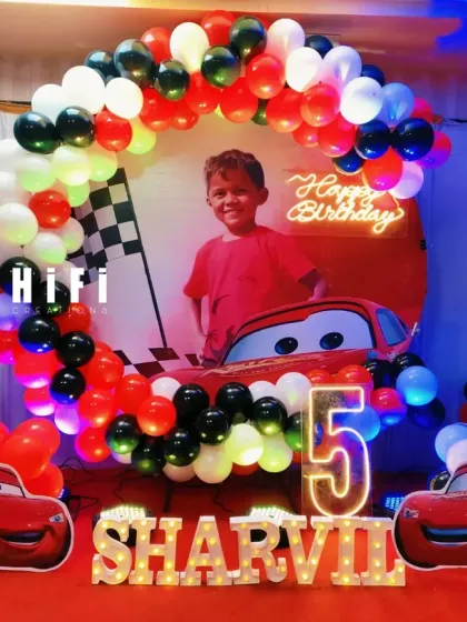 Get ready to race with a Disney Cars themed birthday party. This setup for a 5th birthday includes a circular backdrop with Lightning McQueen, a red, black, and white balloon arch, and multiple character cutouts.