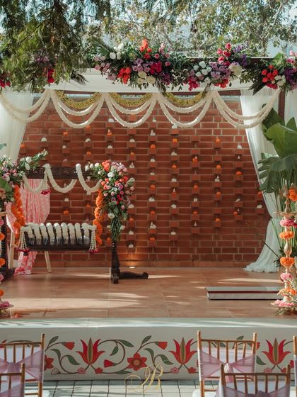 A vibrant stage setup for a naming ceremony against a warm brick wall. The decor features a colorful floral arch over the cradle, with white drapes and traditional banana plants framing the space.