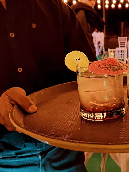 A waiter carries a tray with two expertly prepared cocktails at an outdoor event. Our service is seamless, ensuring your guests are always looked after.