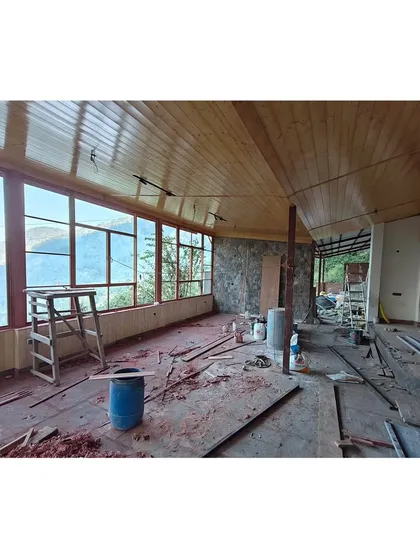 The interior of the pavilion during the construction phase, with tools and materials on the floor. The raw structure of the stone wall and wooden ceiling is visible before final finishing.