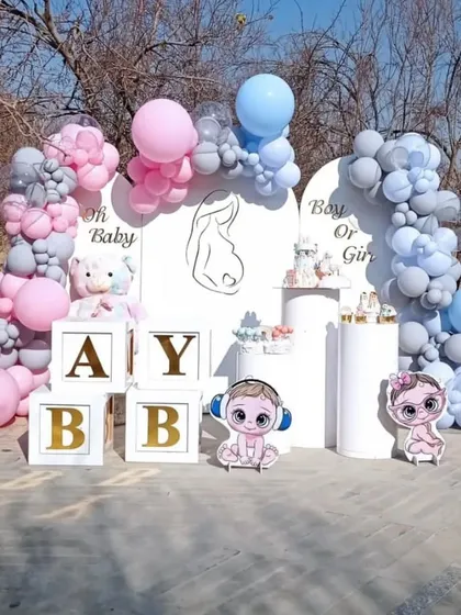 A chic outdoor baby shower decoration with a "Boy or Girl" theme. The setup includes white backdrops, pink and blue balloon arches, "BABY" blocks, and cute baby character cutouts.
