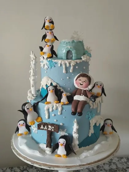 A winter wonderland adventure for a special birthday boy. This two-tiered cake is covered in snowy details, a fondant igloo, and a whole colony of adorable, hand-sculpted penguins. The personalized figurine of the birthday boy makes him the star of this chilly, charming scene.