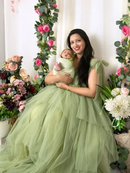 A mother holding her newborn smiles at the camera, surrounded by the lush flowers of our studio setup.