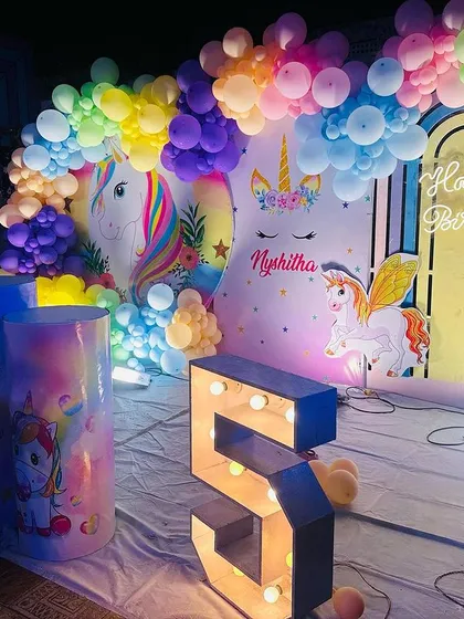 A vibrant unicorn-themed stage decoration. This setup features a large backdrop with unicorns and rainbows, a colorful balloon arch, and a marquee number '5'.
