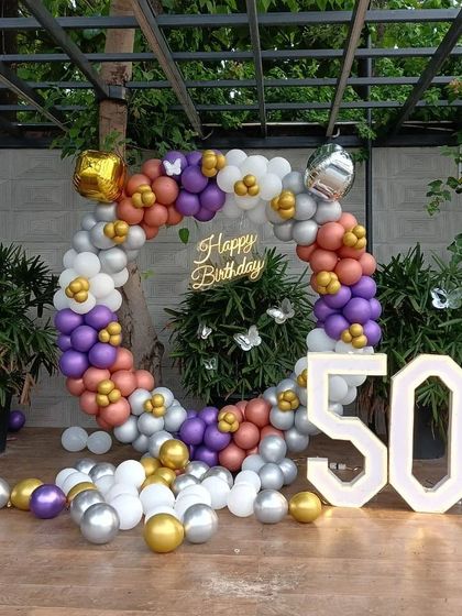 A colorful 50th birthday celebration in a garden setting. This setup features a circular balloon arch in purple, orange, gold, and silver, with large '50' marquee numbers.