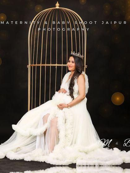 A creative studio portrait featuring a mom-to-be in a white gown, seated in front of a golden birdcage prop. The dark background with bokeh lights adds a magical, whimsical touch.