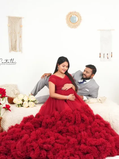 A full-length view of the couple in a boho-chic setup. The wife's red gown contrasts beautifully with the neutral tones of the macrame and cushions.