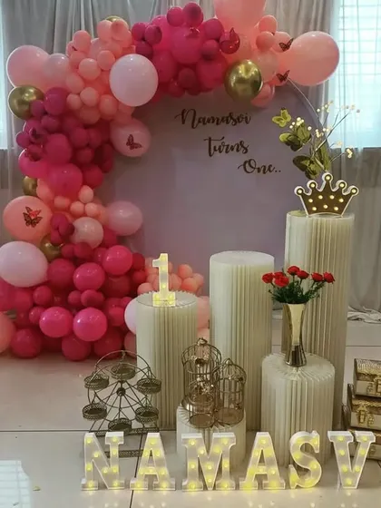 A beautiful pink-themed first birthday decoration at home, with a balloon half-arch, personalized name in lights, and decorative props.