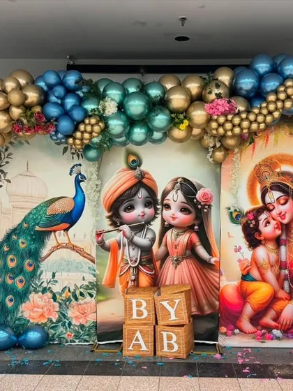 A grand and artistic Krishna-themed backdrop featuring a large peacock and beautiful illustrations. The scene is framed by a magnificent balloon arch in metallic teal and gold.