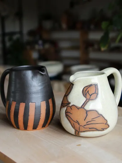 Two pitchers made by students, each with a unique surface decoration. One features a wax-resist pattern, while the other has a delicate, hand-painted lotus design.
