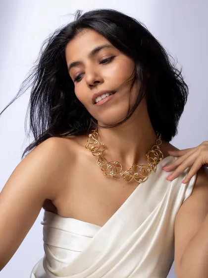 A soft, elegant portrait featuring an abstract gold necklace. The lighting and pose are designed to convey a sense of luxury and artistry.