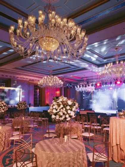 The ballroom setup for the Sangeet, showing the grand chandeliers, elegant table settings, and the impressive stage. I transform blank spaces into opulent environments.